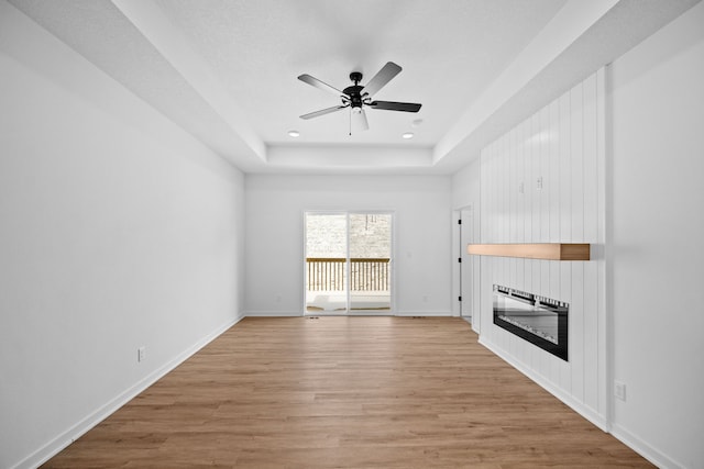 unfurnished living room featuring light wood-type flooring, a large fireplace, a raised ceiling, and ceiling fan