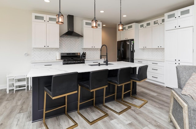 kitchen with a center island with sink, wall chimney range hood, sink, stainless steel gas stove, and black fridge with ice dispenser