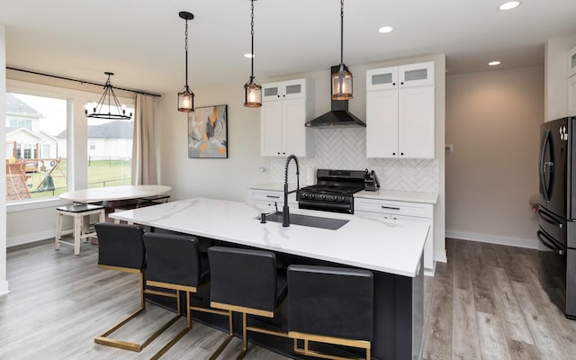 kitchen with a kitchen island with sink, wall chimney range hood, black appliances, sink, and white cabinetry