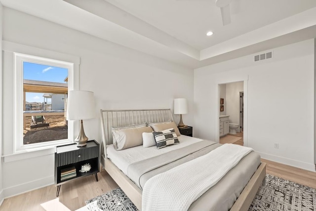 bedroom with a raised ceiling, connected bathroom, ceiling fan, and light wood-type flooring