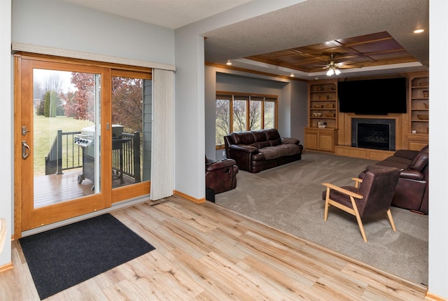 living room featuring built in features, a fireplace, ceiling fan, a raised ceiling, and light hardwood / wood-style flooring