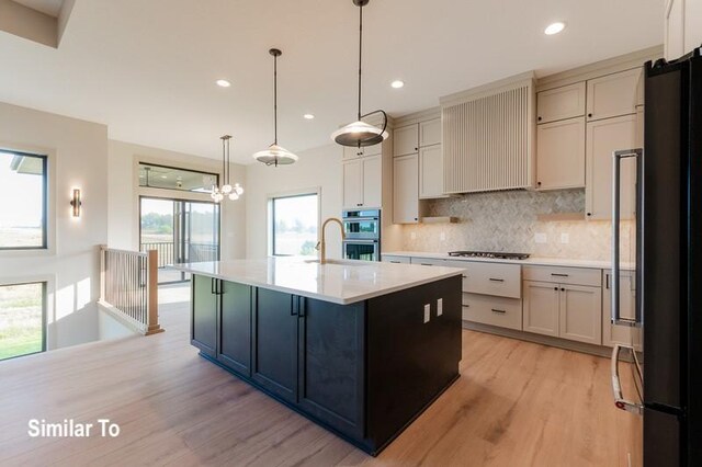 kitchen featuring premium range hood, stainless steel appliances, a kitchen island with sink, decorative light fixtures, and light hardwood / wood-style flooring