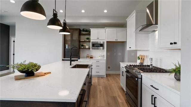 kitchen with white cabinetry, hanging light fixtures, wall chimney range hood, an island with sink, and appliances with stainless steel finishes