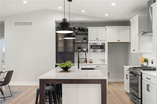 kitchen featuring wall chimney exhaust hood, pendant lighting, a center island with sink, white cabinets, and appliances with stainless steel finishes