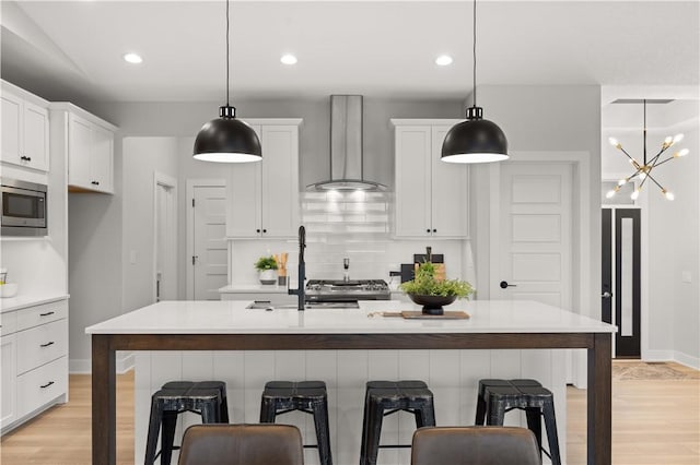 kitchen featuring backsplash, wall chimney exhaust hood, stainless steel microwave, a kitchen island with sink, and hanging light fixtures