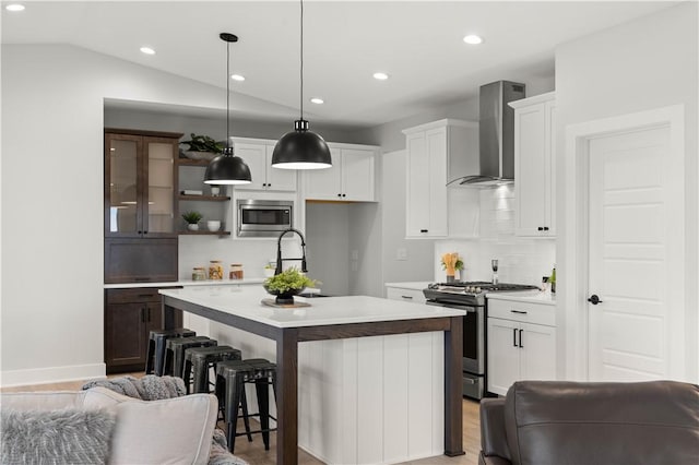 kitchen featuring pendant lighting, lofted ceiling, wall chimney range hood, an island with sink, and appliances with stainless steel finishes