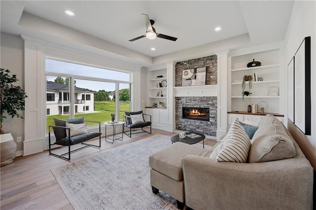 living room with a stone fireplace, ceiling fan, a tray ceiling, light hardwood / wood-style floors, and built in shelves