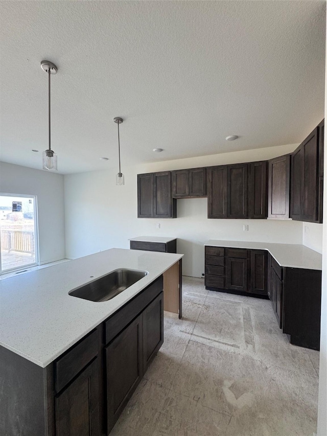 kitchen featuring a kitchen island with sink, a textured ceiling, dark brown cabinets, pendant lighting, and a sink