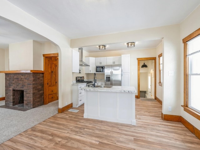 kitchen featuring white cabinets, appliances with stainless steel finishes, a brick fireplace, and wall chimney range hood