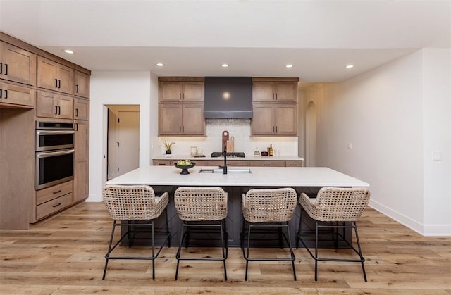 kitchen with double oven, an island with sink, wall chimney exhaust hood, and light wood-type flooring