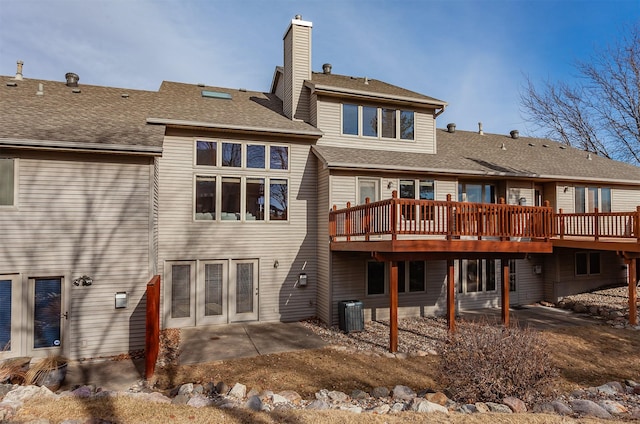 rear view of property with a patio, central AC unit, roof with shingles, a wooden deck, and a chimney