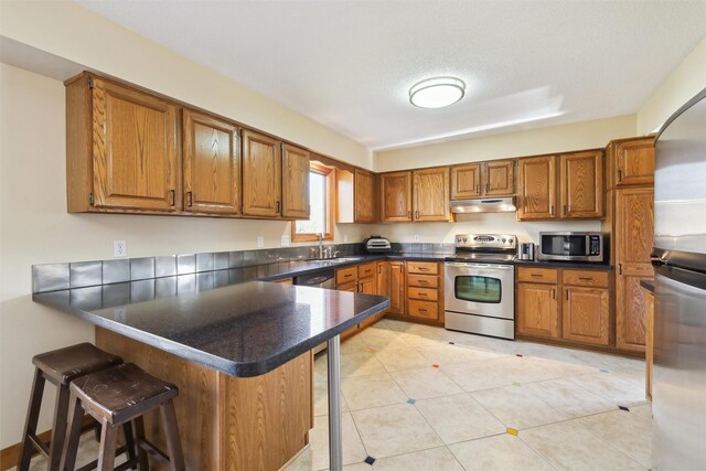 kitchen with under cabinet range hood, a breakfast bar area, brown cabinets, appliances with stainless steel finishes, and a peninsula