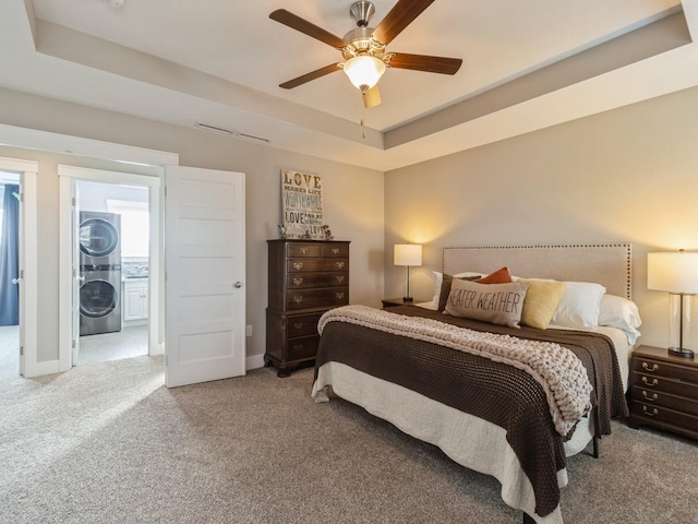 carpeted bedroom with a tray ceiling, connected bathroom, ceiling fan, and stacked washer / drying machine