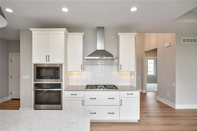 kitchen with appliances with stainless steel finishes, wall chimney range hood, light hardwood / wood-style floors, decorative backsplash, and white cabinets