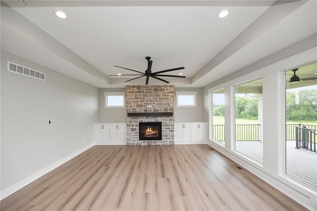 unfurnished living room with a tray ceiling, a stone fireplace, light hardwood / wood-style floors, and ceiling fan