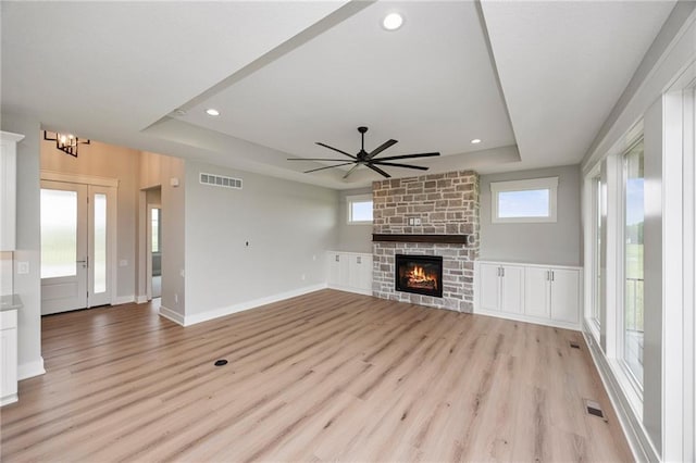 unfurnished living room with ceiling fan, a tray ceiling, a stone fireplace, and light wood-type flooring