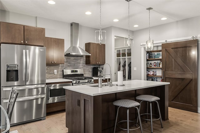 kitchen featuring stainless steel appliances, a kitchen island with sink, sink, wall chimney range hood, and a barn door