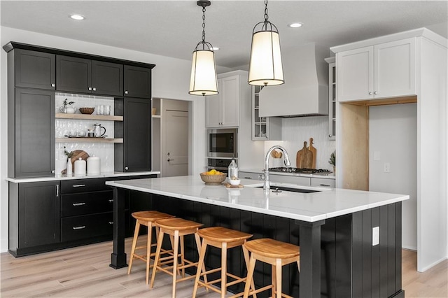 kitchen with built in microwave, wall chimney range hood, white cabinets, and a center island with sink