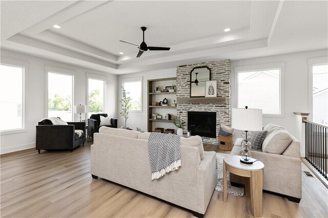 living room with ceiling fan, built in features, a tray ceiling, a fireplace, and light wood-type flooring