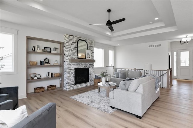 living room featuring a tray ceiling, built in features, ceiling fan, a fireplace, and light hardwood / wood-style floors