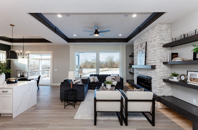 living room with ornamental molding, ceiling fan with notable chandelier, a tray ceiling, light hardwood / wood-style flooring, and a stone fireplace