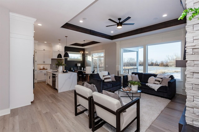 living room with a wealth of natural light, ornamental molding, ceiling fan with notable chandelier, a raised ceiling, and light hardwood / wood-style flooring