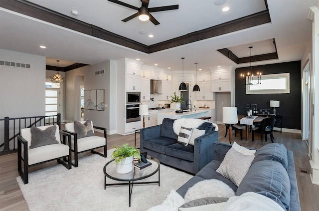 living room featuring ornamental molding, ceiling fan with notable chandelier, a tray ceiling, sink, and light hardwood / wood-style flooring