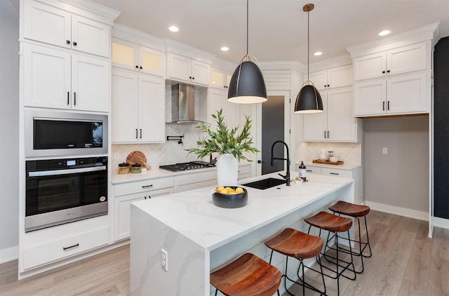 kitchen with wall chimney range hood, appliances with stainless steel finishes, a kitchen island with sink, and sink