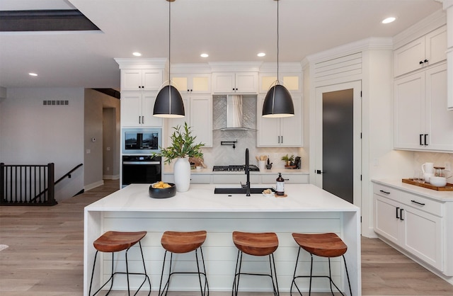 kitchen with tasteful backsplash, a kitchen island with sink, white cabinets, and wall chimney range hood