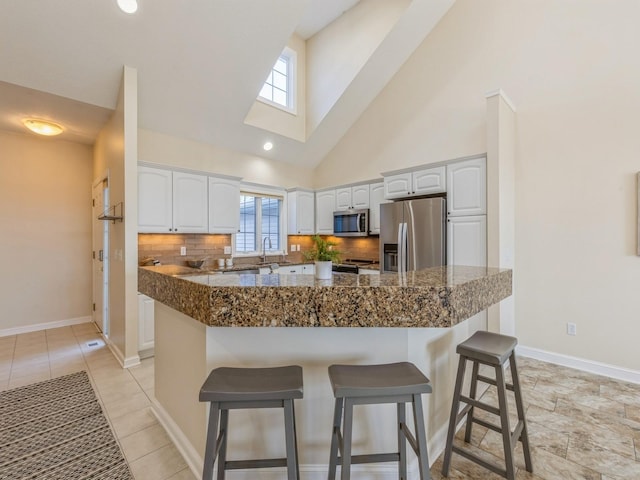 kitchen with high vaulted ceiling, kitchen peninsula, appliances with stainless steel finishes, a kitchen bar, and white cabinetry