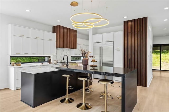 kitchen featuring a breakfast bar area, hanging light fixtures, stainless steel built in refrigerator, an island with sink, and decorative backsplash