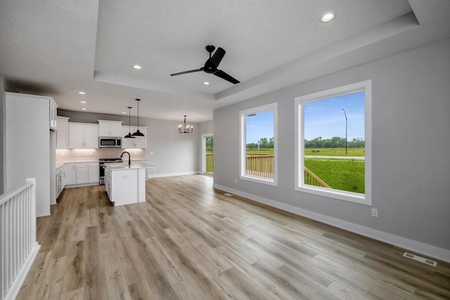 unfurnished living room with a raised ceiling, plenty of natural light, light hardwood / wood-style flooring, and ceiling fan with notable chandelier