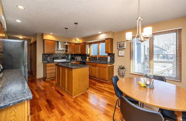 kitchen featuring stove, stainless steel refrigerator, hanging light fixtures, a kitchen island, and wall chimney range hood