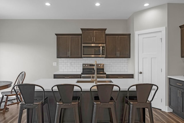 kitchen with sink, an island with sink, decorative backsplash, dark hardwood / wood-style flooring, and appliances with stainless steel finishes