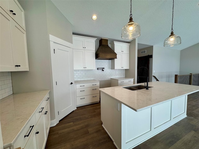 kitchen featuring custom exhaust hood, hanging light fixtures, an island with sink, white cabinets, and sink