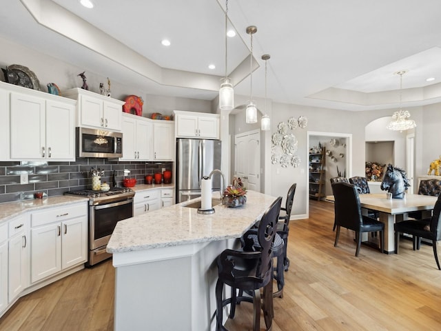 kitchen featuring hanging light fixtures, stainless steel appliances, a raised ceiling, a kitchen island with sink, and white cabinetry