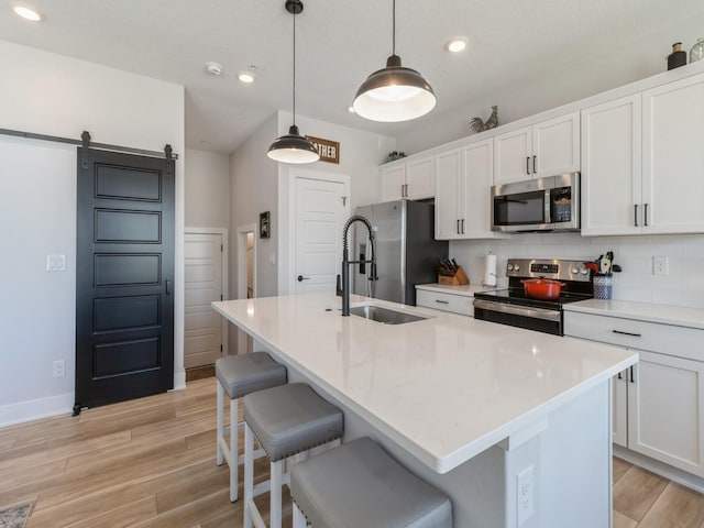 kitchen with white cabinets, hanging light fixtures, a barn door, a center island with sink, and appliances with stainless steel finishes