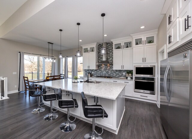 kitchen with wall chimney exhaust hood, white cabinetry, stainless steel appliances, light stone counters, and a center island with sink