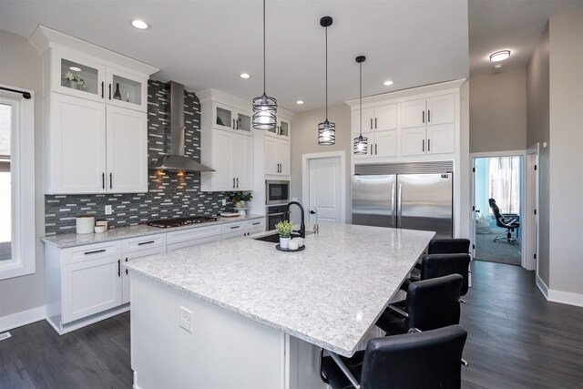 kitchen featuring white cabinetry, wall chimney range hood, a center island with sink, and built in appliances