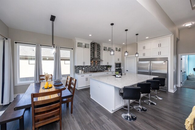 kitchen with white cabinetry, wall chimney range hood, built in appliances, hanging light fixtures, and a kitchen island with sink