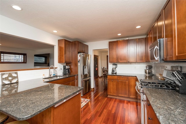 kitchen with kitchen peninsula, sink, dark wood-type flooring, appliances with stainless steel finishes, and dark stone counters