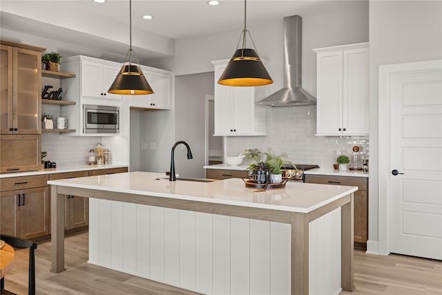 kitchen featuring stainless steel microwave, wall chimney exhaust hood, hanging light fixtures, and a kitchen island with sink