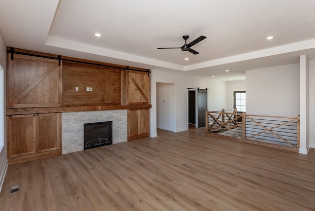 unfurnished living room featuring ceiling fan, a barn door, a tile fireplace, a raised ceiling, and wood-type flooring