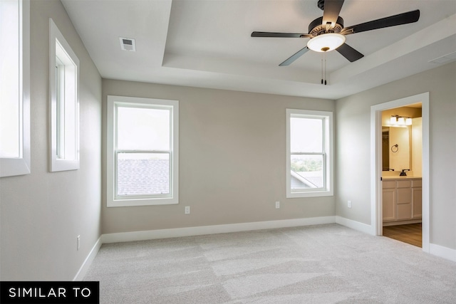 unfurnished bedroom featuring ensuite bath, light colored carpet, ceiling fan, and a raised ceiling