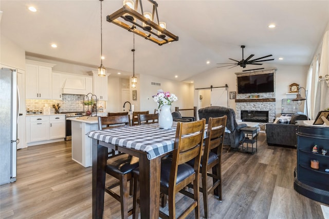 dining room with a barn door, a stone fireplace, ceiling fan, wood-type flooring, and vaulted ceiling