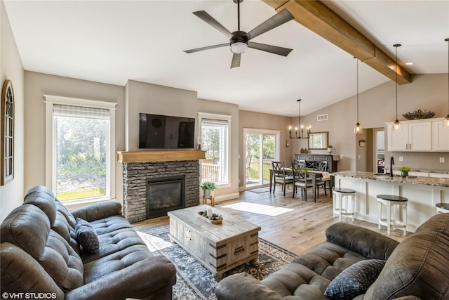 living room featuring ceiling fan with notable chandelier, light hardwood / wood-style floors, a stone fireplace, and lofted ceiling with beams