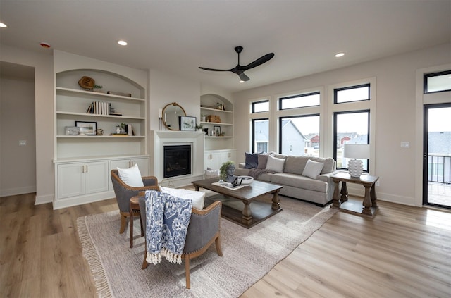 living room featuring a ceiling fan, a glass covered fireplace, built in shelves, and light wood-type flooring