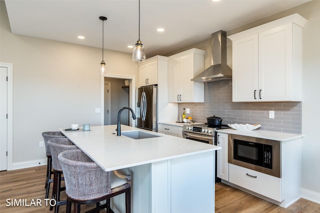 kitchen featuring decorative light fixtures, appliances with stainless steel finishes, wall chimney range hood, and a center island with sink