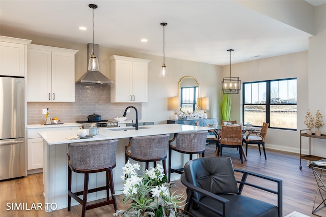 kitchen with a center island with sink, sink, stainless steel refrigerator, white cabinetry, and wall chimney exhaust hood