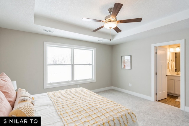 bedroom with a textured ceiling, ensuite bathroom, ceiling fan, light colored carpet, and a tray ceiling